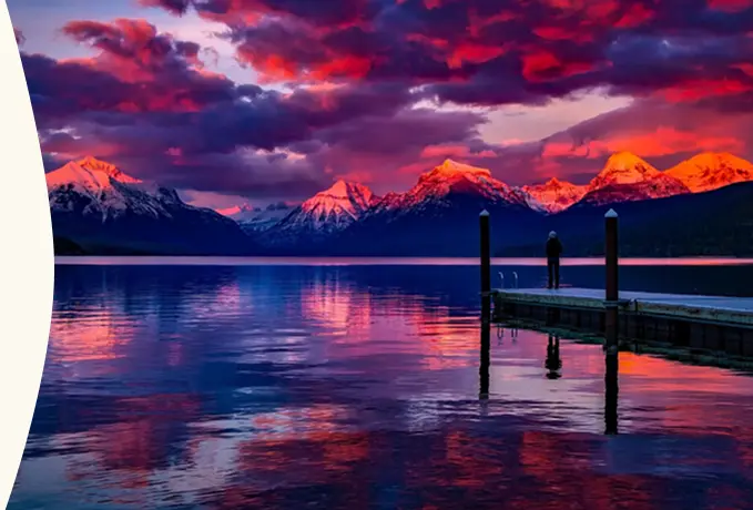 Stunning view of mountains at sunset, with colorful reflections in the calm lake and a lone person standing on a dock.