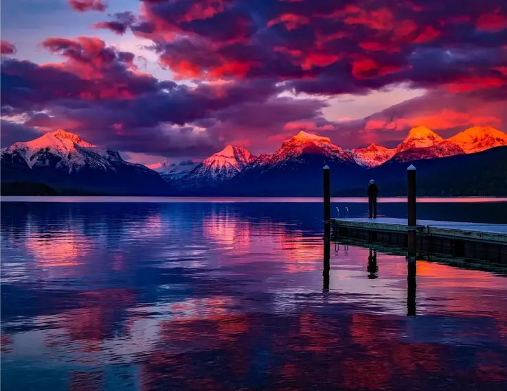 Stunning view of mountains at sunset, with colorful reflections in the calm lake and a lone person standing on a dock.