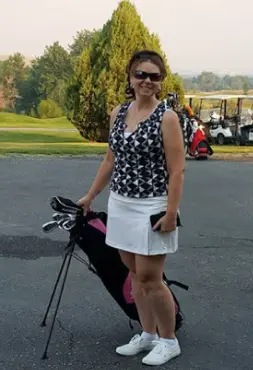 Woman holding a golf club, wearing a patterned top, white skirt, and sneakers on a golf course.