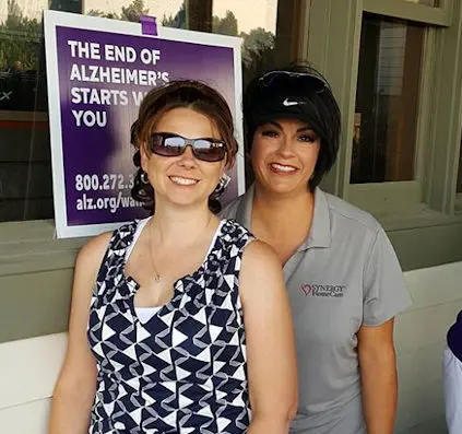 Two women standing together in front of an Alzheimer's awareness poster, both smiling and wearing casual attire.