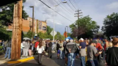 A crowd of people gathered on a city street, with buildings and power lines in the background.