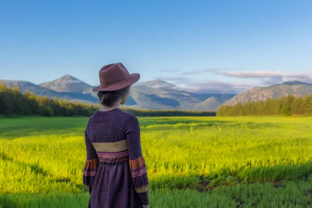 Person wearing a hat standing in a green field with mountains in the background.