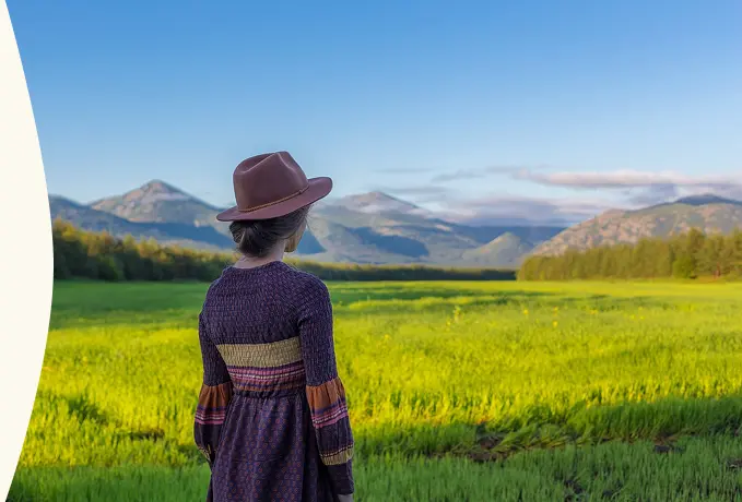 Person wearing a hat standing in a green field with mountains in the background.