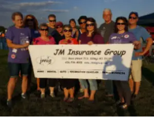 Group of people holding a 'JM Insurance Group' banner at a Relay for Life event, wearing purple shirts.